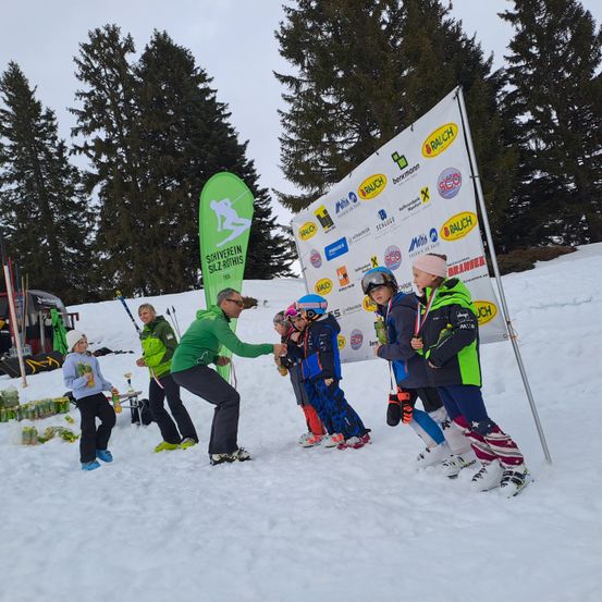 Eine Gruppe von Kindern und Erwachsenen in Winterkleidung steht im Schnee, einige erhalten Medaillen. Ein erwachsener Mann überreicht einem Kind eine Medaille. Dahinter steht ein Banner mit Logos und Text.
