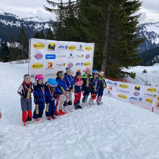 Eine Gruppe junger Skifahrer mit Medaillen um den Hals steht auf einem verschneiten Hang. Sie stehen vor einem Banner mit verschiedenen Sponsorenlogos. Im Hintergrund sind verschneite Berge zu sehen.