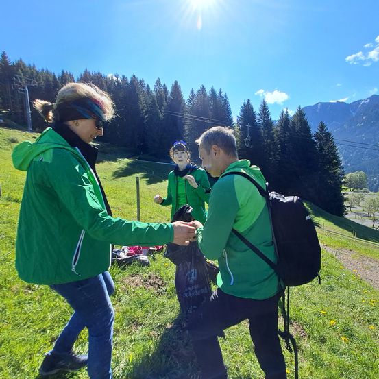 Drei Personen in grünen Jacken stehen auf einem grünen Hügel unter einem hellen Himmel. Eine Frau lächelt, während sie eine Plastiktüte hält. Ein Mann kniet, um einer anderen Person eine Plastiktüte zu geben.