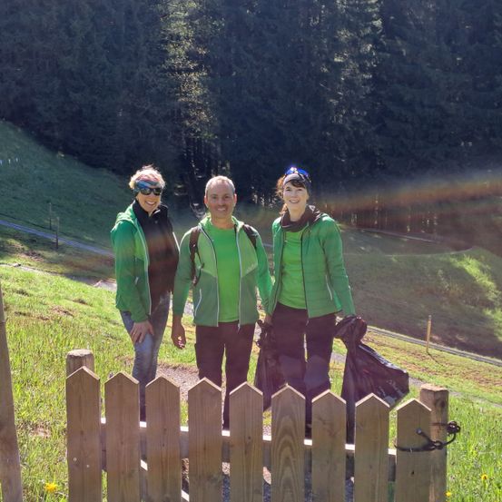 Drei Personen, gekleidet in grünen Jacken, posieren für ein Foto in einem Grasbereich mit einem Holzzaun, in der Nähe eines Berges mit Bäumen.