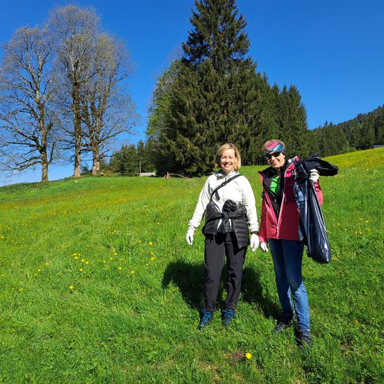 Zwei Frauen stehen in einer üppigen, grünen Wiese mit einem Hintergrund aus Bäumen und Bergen. Sie tragen Handschuhe und tragen Taschen. Eine Frau trägt einen Hut und eine Sonnenbrille.
