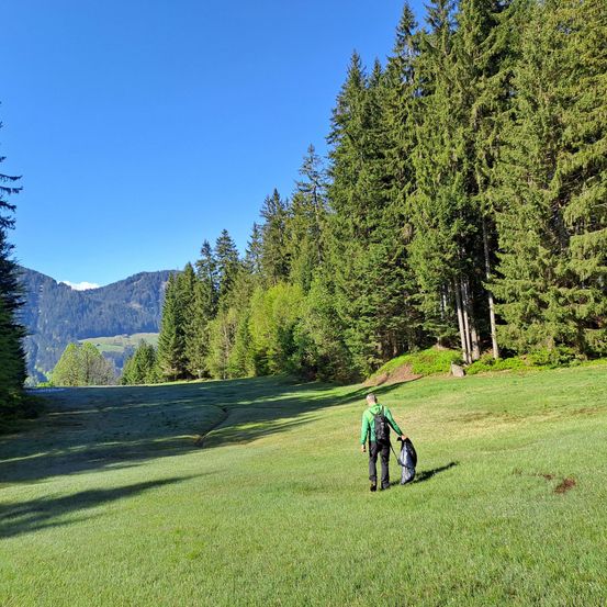 Ein Mann in einem grünen Jackett geht auf einem Grasfeld mit einem Golfbag. Hohe Bäume umgeben das Gebiet, und Berge sind in der Ferne sichtbar.