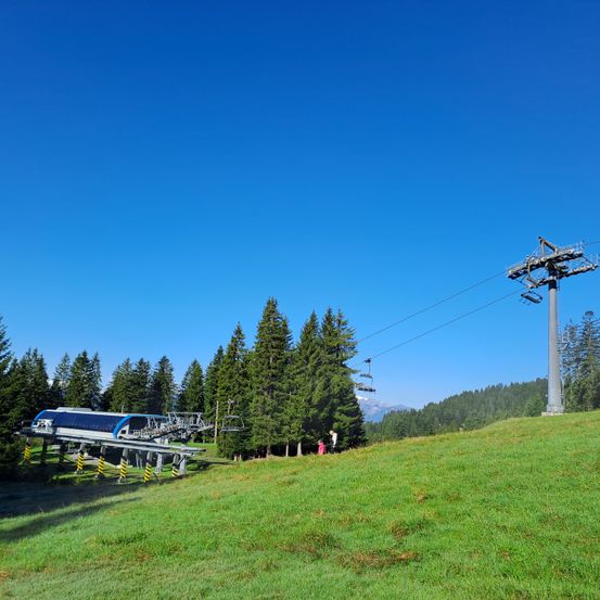 Eine Skiliftstation steht auf einem grünen Hügel bei klarem blauem Himmel. Hohe Bäume umgeben das Gebiet, und einige Leute sind in der Nähe des Lifts.