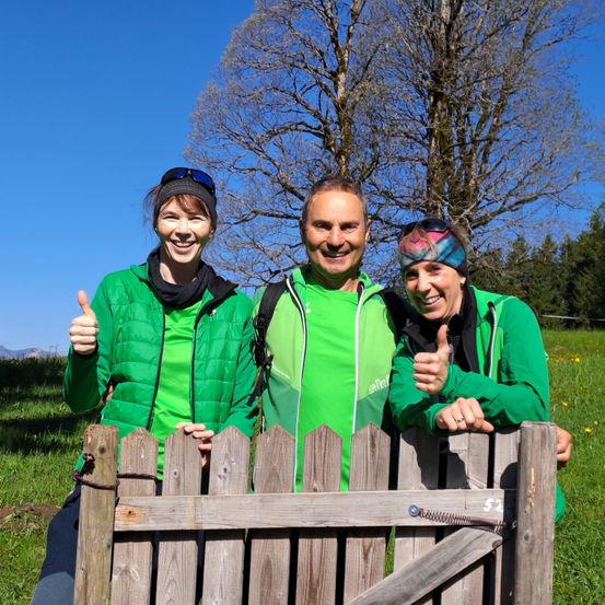 Drei Menschen in grünen Jacken und Stirnbändern, die lächeln und Daumen hoch halten, während sie hinter einem Holzzaun in einem Grasfeld mit Bäumen im Hintergrund stehen.