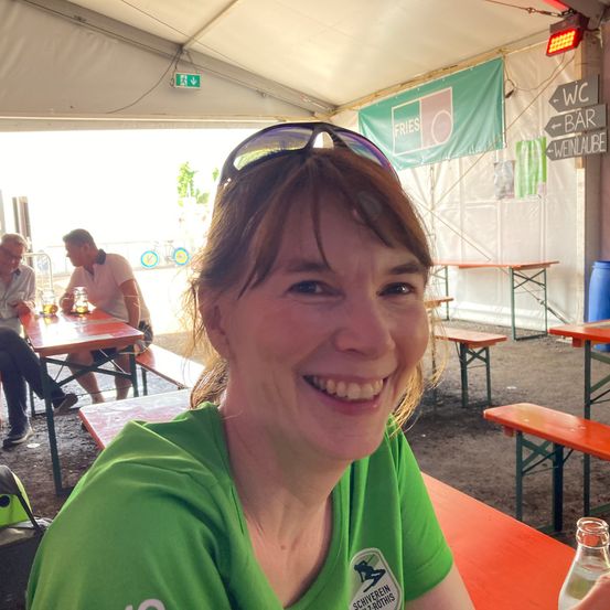 Eine Frau in einem grünen Shirt mit Logo sitzt an einem Tisch in einem Zelt. Andere Leute sitzen an anderen Tischen im Hintergrund. Es gibt Schilder für WC und Bar.