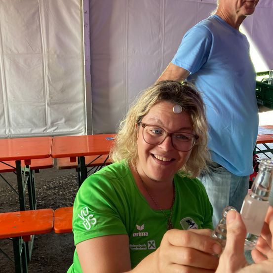 Eine Frau mit Brille lächelt, während sie eine Flasche hält und ein Abzeichen auf ihrer Stirn zeigt. Ein Mann in einem blauen T-Shirt steht hinter ihr. Sie befinden sich in einem Zelt mit orangefarbenen Tischen und weißen Wänden.