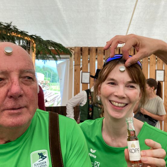 Ein Mann und eine Frau tragen grüne T-Shirts und stehen nebeneinander. Der Mann hat eine Münze auf dem Kopf, während die Frau eine Flasche in der Hand hält. Hinter ihnen stehen Menschen in einem Raum mit Holzwänden und einem Fenster.