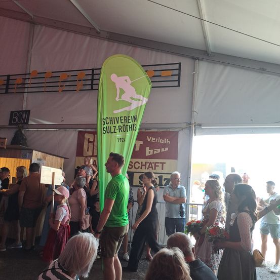 Eine Menschenmenge versammelt sich unter einem Zelt, ein Mann in einem grünen T-Shirt sticht hervor, ein großer Banner hinter ihm trägt die Aufschrift 'Schiverein Sulz-Rothis'. Menschen stehen und beobachten, einige halten Blumen.