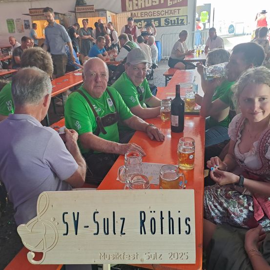 Eine Gruppe von Menschen in grünen Shirts sitzt an einem Tisch im Freien und trinkt Bier und Wein. Ein Holzschild mit dem Text 'SV-Sulz Rotis Musikfest Sulz 2025' steht vor ihnen.