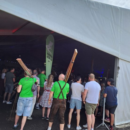 In einem großen Zelt stehen mehrere Menschen in einer Schlange. Sie tragen alle grüne T-Shirts, Shorts und Mützen. Einige von ihnen haben Holzstöcke auf den Schultern. Das Zelt ist weiß und auf der linken Seite befindet sich ein grüner Banner mit weißen Text.