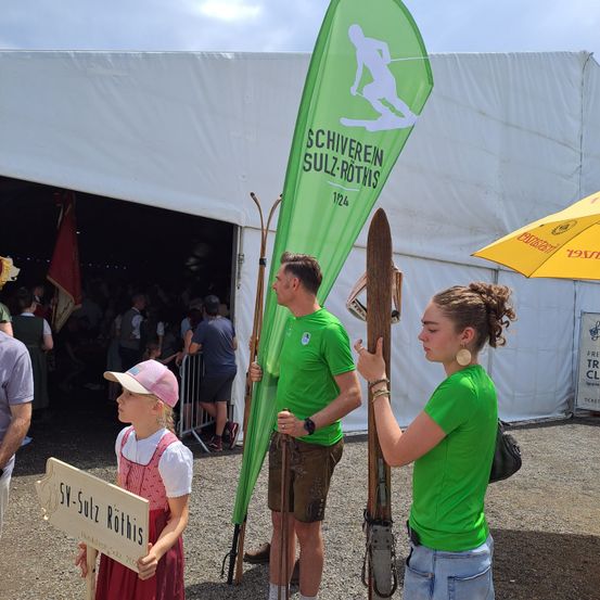 Eine Gruppe von Menschen in grünen Shirts steht vor einem weißen Zelt mit einem grünen Banner, das 'Schiferien Sulz-Rotis' steht. Ein Mädchen in einer rosa Mütze hält ein Schild, und ein Mann neben ihr hält einen Skistock. Ein Regenschirm und Flaggen sind hinter ihnen.