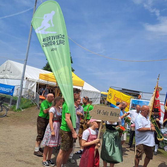 Eine Gruppe von Menschen in traditioneller Kleidung steht in einem Außenbereich, möglicherweise für ein Festival. Ein junges Mädchen hält ein Schild mit der Aufschrift 'SV-Sulz Rothis'. Dahinter ist ein grüner Banner mit weißem Text zu sehen. Das Gebiet ist von Zelten und Fahrrädern umgeben.