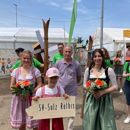 Eine Gruppe von Menschen in traditioneller Kleidung posiert für ein Foto bei einer Outdoor-Veranstaltung, hält Blumen und Schilder in der Hand, mit Zelten und Windmühlen im Hintergrund.