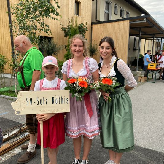 Eine Gruppe von Menschen posiert für ein Foto vor einem Gebäude. Ein Mädchen hält ein Schild und Blumen, während andere traditionelle Kleidung tragen. Ein älterer Mann steht hinter ihnen.