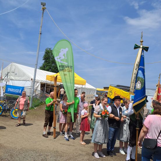 Eine Gruppe von Menschen versammelt sich draußen, einige in traditioneller Kleidung, in der Nähe von Zelten und Bannern. Ein Mann hält ein Schild und eine Frau mit Blumen steht neben ihm.
