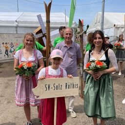 Zwei Frauen und ein Mädchen mit Blumen und ein Junge mit einem Schild bei einem Musikfestival, stehen vor einem weißen Zelt.