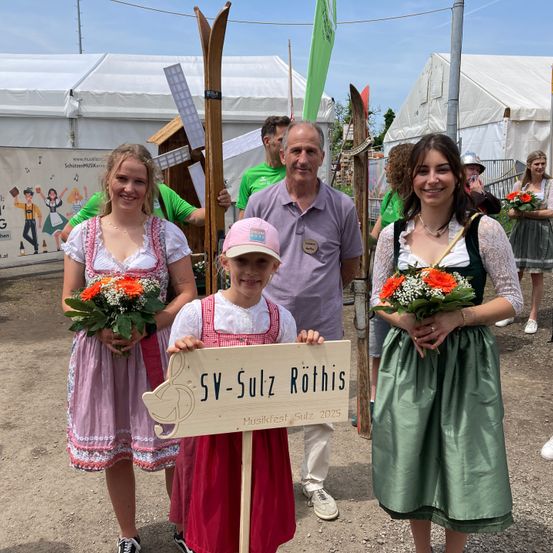 Zwei Frauen und ein Mädchen mit Blumen und ein Junge mit einem Schild bei einem Musikfestival, stehen vor einem weißen Zelt.