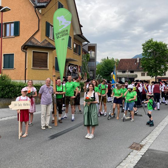 Eine Gruppe von Menschen in grünen Shirts und Kleidern marschiert in einem Umzug. Ein Mädchen mit einem Schild und eine Frau mit Blumen sind vorne. Ein Banner ist an einem Pfahl dahinter befestigt. Gebäude, Bäume und Berge sind im Hintergrund.