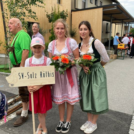 Drei Frauen und ein junges Mädchen stehen vor einem Holzgebäude. Das Mädchen hält ein Schild mit der Aufschrift 'SV-Sulz Rothis Musikfest Sulz 2025'. Sie halten alle Blumensträuße. Ein älterer Mann steht hinter ihnen.
