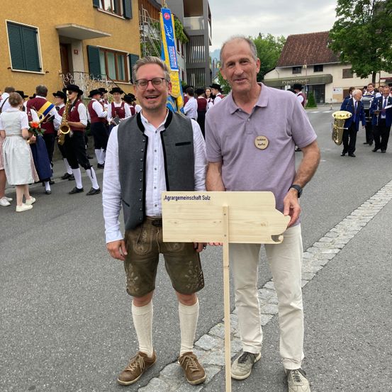 Zwei Männer stehen während eines Festes in einer Straße und lächeln. Einer hält ein Holzschild. Eine Band spielt hinter ihnen.