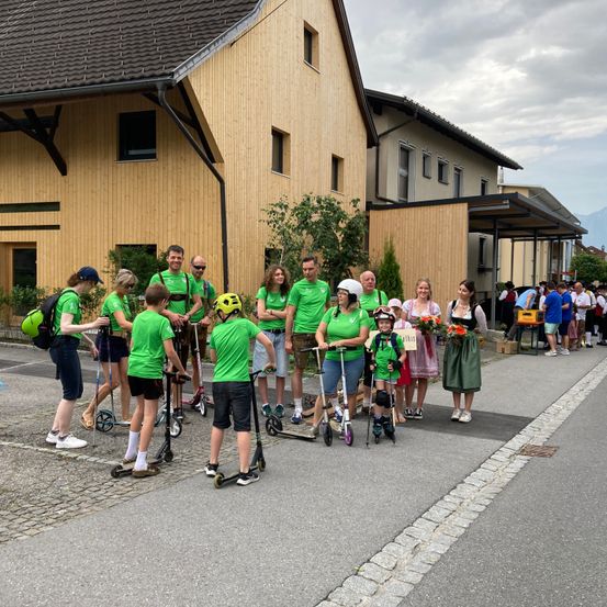 Eine Gruppe von Menschen in grünen Shirts und Helmen steht in einer Reihe mit Rollern vor einem Gebäude bei bewölktem Himmel.