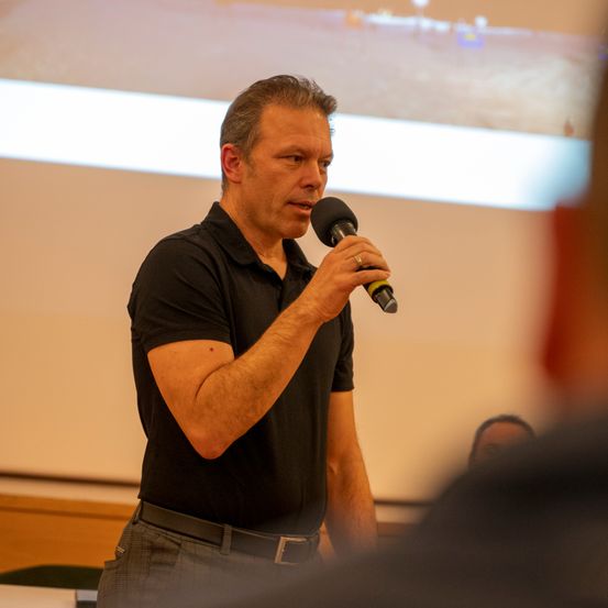 Ein Mann spricht in ein Mikrofon vor einer Projektionsleinwand, die eine Baustelle zeigt. Er trägt ein schwarzes Hemd und einen Ring an seinem Finger.