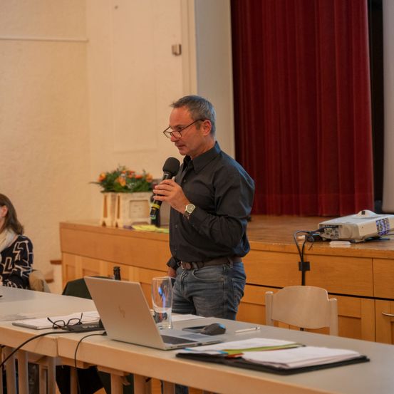 Ein Mann mit Brille und einer Uhr steht in einem Konferenzraum an einem Tisch und hält ein Mikrofon. Ein Laptop, Papiere und ein Glas Wasser stehen auf dem Tisch. Ein Beamer ist auf der Bühne hinter ihm, und eine Frau sitzt an einem anderen Tisch.