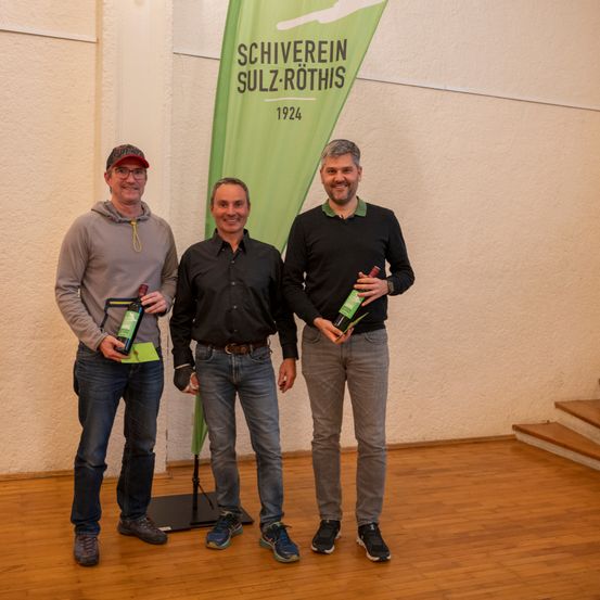 Drei Männer stehen nebeneinander, halten Flaschen in der Hand, lächeln und posieren für ein Foto vor einem Banner mit dem Text 'Schiverein Sulz-Rothis 1924'.