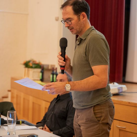 Ein Mann spricht in ein Mikrofon und hält ein Papier in der Hand, während ein anderer Mann im Hintergrund sitzt. Dahinter steht ein Podest mit Blumen, Flaschen und einem Projektor.