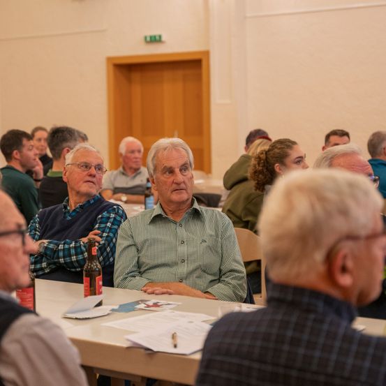 Eine Gruppe älterer Männer und Frauen sitzt an einem Tisch, einige mit Brille. Papiere und Flaschen sind auf dem Tisch. Eine Tür ist im Hintergrund zu sehen.