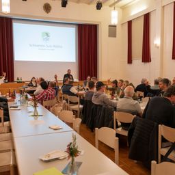 Eine Gruppe von Menschen sitzt um Tische in einem Raum herum und schaut sich eine Präsentation auf einer Projektionsleinwand an. Auf den Tischen stehen Flaschen, Teller und Blumen. Einige Leute heben ihre Hände.