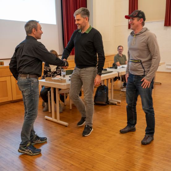 Drei Männer stehen in einem Raum, zwei schütteln Hände, während der dritte zusieht. Ein Schreibtisch mit Gegenständen steht hinter ihnen, und Menschen sitzen an einem anderen Tisch. Eine Projektionsleinwand ist an der Wand.