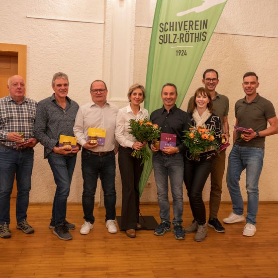 Eine Gruppe von Menschen mit Büchern und Blumen, die vor einer Fahne mit 'SCHIVEREI SULZ-RUTHIS 1924' stehen.