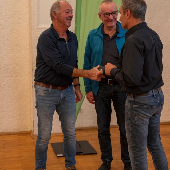 Drei Männer stehen lächelnd zusammen drinnen. Der linke Mann schüttelt dem mittleren die Hand. Der mittlere trägt eine Brille und eine blaue Jacke. Der rechte trägt ein schwarzes Hemd. Hinter ihnen ist eine grüne Gardine.