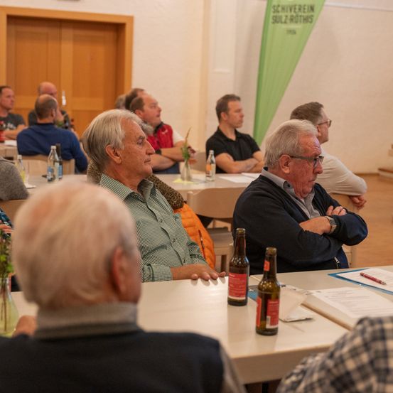 Eine Gruppe älterer Männer sitzt um einen Tisch herum, manche mit verschränkten Armen, in einer Besprechung. Flaschen und Papiere liegen auf dem Tisch. Ein Banner ist im Hintergrund.