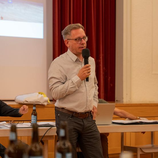 Ein Mann steht vor einem Tisch und hält ein Mikrofon in der Hand, wahrscheinlich hält er eine Präsentation. Hinter ihm zeigt eine Projektionsleinwand eine Folie.