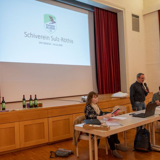 Eine Frau liest aus einem Buch, während sie an einem Tisch in einem Besprechungsraum sitzt. Ein Mann spricht in ein Mikrofon, und eine Präsentation wird auf einer Projektionsleinwand gezeigt.