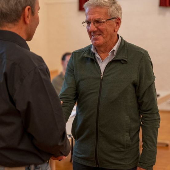 Ein älterer Mann mit Brille und einer grünen Jacke schüttelt einem anderen Mann in einem schwarzen Hemd die Hand. Hinter ihnen sitzt ein Mann auf einem Stuhl. Der Raum hat Holzböden und weiße Wände.