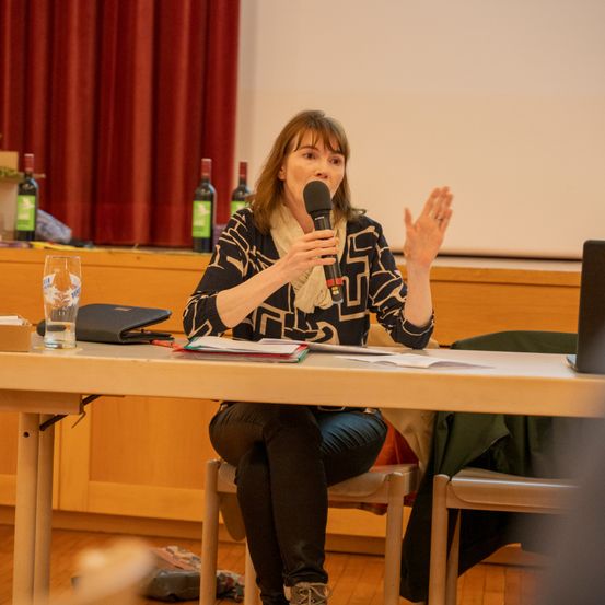 Eine Frau sitzt an einem Tisch, hält ein Mikrofon und gestikuliert, möglicherweise während einer Konferenz. Hinter ihr sind Weinflaschen und ein roter Vorhang zu sehen.