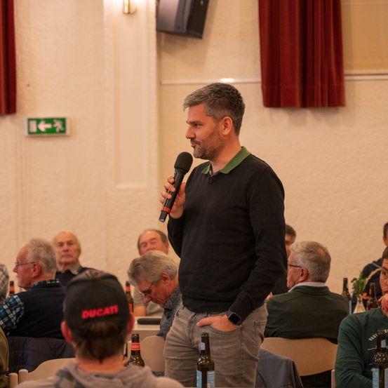 Ein Mann steht vor einer Menschenmenge und hält ein Mikrofon. Er trägt ein schwarzes Langarmshirt und graue Hose. Hinter ihm sitzen Menschen in Stühlen, einige mit Flaschen. Eine rote Gardine hängt an der Wand.