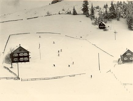 Ein Schwarz-Weiß-Luftbild einer verschneiten Landschaft mit mehreren Skifahrern. Es gibt zwei Gebäude, eines mit einer Flagge, und Bäume in der Umgebung.