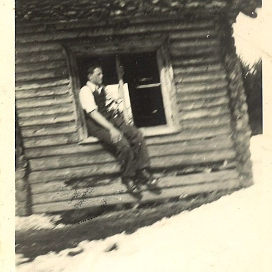 Ein Schwarz-Weiß-Foto zeigt einen Mann, der aus einem Fenster eines Blockhauses lehnt. Er trägt ein Hemd und eine Latzhose.