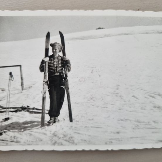 Ein Schwarz-Weiß-Foto zeigt eine Frau, die im Schnee mit Skiern auf den Schultern steht. Sie ist warm gekleidet, und im Hintergrund befindet sich ein Zaun.