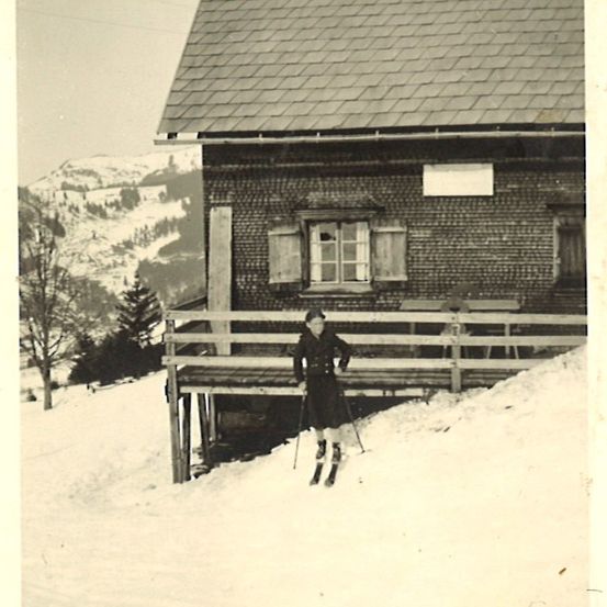 Ein Schwarz-Weiß-Foto einer Frau, die vor einem Haus mit Veranda und Bergen im Hintergrund auf Schnee ski fährt.