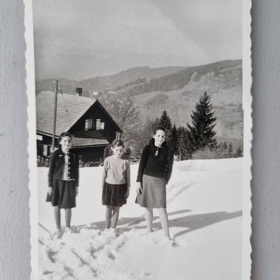 Drei Mädchen stehen im Schnee vor einem Chalet mit einer Bergkette im Hintergrund.