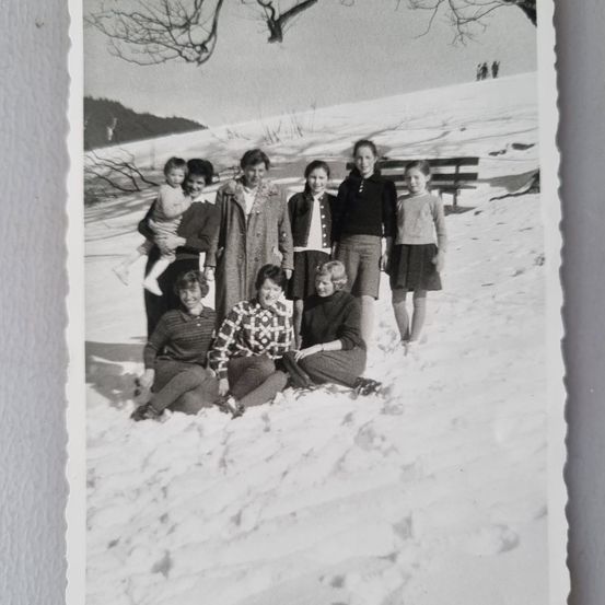Ein Vintage-Schwarz-Weiß-Foto einer Familie auf einem verschneiten Hügel. Sie sind alle warm gekleidet. Hinter ihnen steht eine Bank und einige Bäume. In der Ferne gehen Leute spazieren.