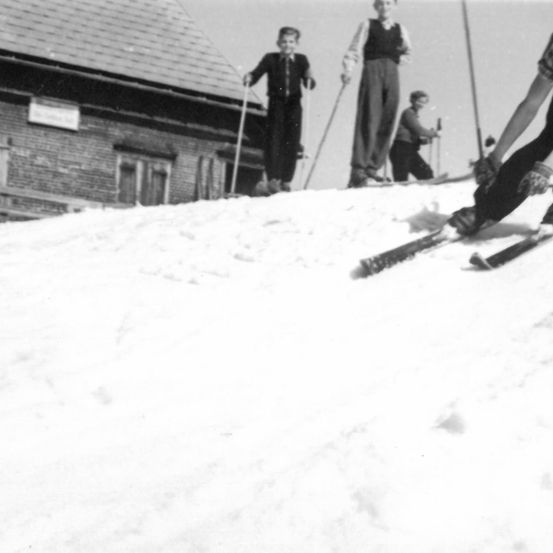 Ein Schwarz-Weiß-Bild zeigt mehrere Kinder, die vor einem Backsteingebäude mit geneigtem Dach Ski fahren. Sie halten alle Skistöcke in den Händen.
