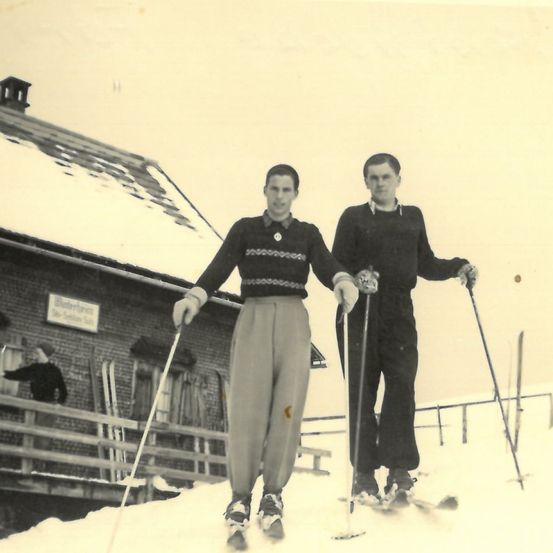 Zwei Männer stehen auf einem verschneiten Hang und tragen Vintage-Skiausrüstung. Einer hat einen gestrickten Pullover, während der andere ein Hemd mit Kragen trägt. Hinter ihnen steht ein Gebäude mit einem Schild und einer weiteren Person auf einem Balkon.