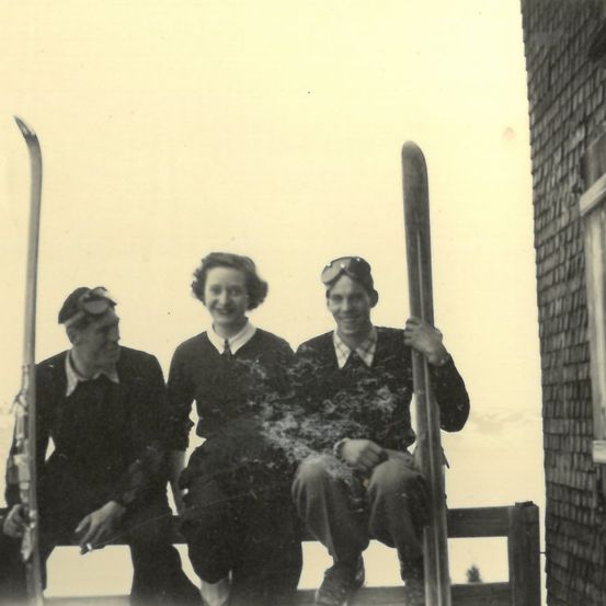 Drei Personen, zwei Männer und eine Frau, sitzen auf einer Holzbank. Sie halten alle Skistöcke. Der Mann rechts trägt eine Mütze mit Brille. Die Frau trägt einen weißen Kragen. Der Hintergrund ist unscharf.