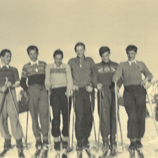 Ein Schwarz-Weiß-Foto von fünf jungen Männern in Skiausrüstung, die in einer Reihe im Schnee stehen.